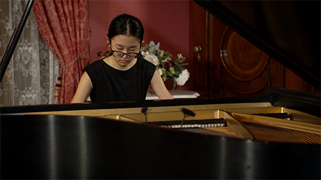 A young woman in a black dress sits playing at a grand piano.