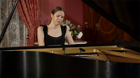 A young woman in a black dress sits playing at a grand piano.