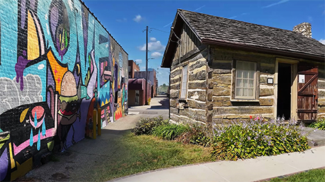 A modern alley mural juxtaposed in a side-by-side composition with a log cabin at a historical village.
