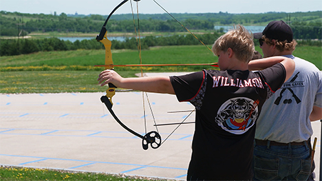 Two males stand next to each other. The one of the left draws his bow and arrow and points it towards a target.