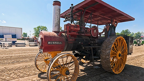 A large antique thresher drives by on the dirt.