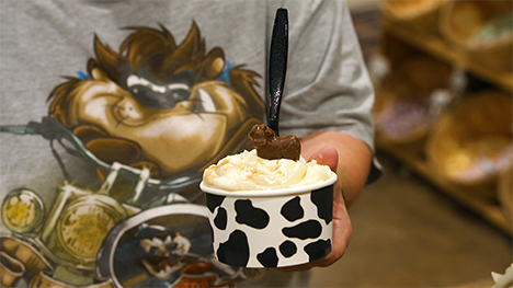 A person holds a cup of ice cream in a cow pattern cup. A spoon and chocolate cow stand vertically from the top of the ice cream.