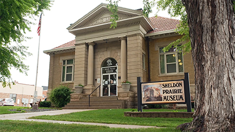 An outside view of the Sheldon Prairie Museum. A flagpole on the left. A sign and tree on the right.