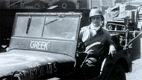 George Soumas sits in a military jeep with a helmet on. The word "GREEK" is under the front windshield.
