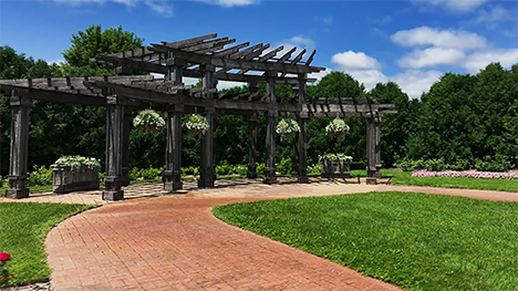 A wooden structure rises up from the grounds of the Cedar Valley Arboretum and Botanic Gardens. Plants hang symmetrically from the structure. A brick pathway curves past the front of the structure.