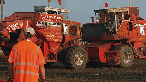 Two large, red combines bash head-on into each other as a man wearing a ball cap and safety shirt in the foreground watches on.