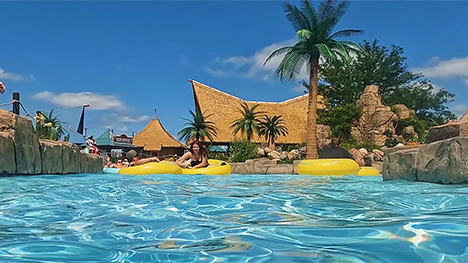 A water, surface level view of inner tubers on clear water at a tropical-themed water park. Palm trees, thatch roofs and yellow inner tubes are viewable.