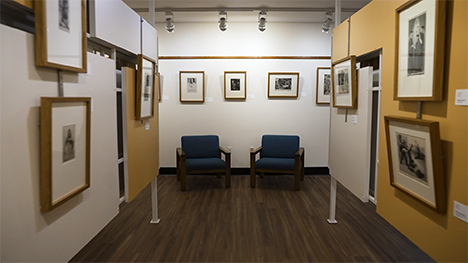 An interior, symmetrical view down an isle of an art gallery. Artwork hangs from all walls. Two blue cushioned chairs sit symmetrically next to each other on a wooden floor against the wall.