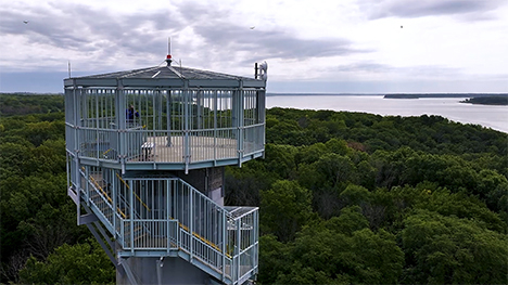 High above the treetops, a man stands at the top of a caged, metal observation tower and shoots video out towards nearby Red Rock Lake.