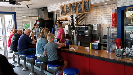 Five people/customers of various ages sit on stools at a restaurant counter. A waitress assists the customers with counterservice.