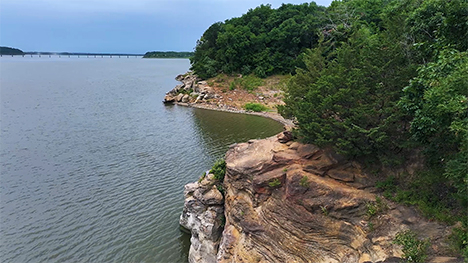 The curvy and rocky cliffs of Red Rock Lake's shoreline. A long vehicle bridge is seen in in the distance. Lush green trees adorn the top of the rocky cliffs.