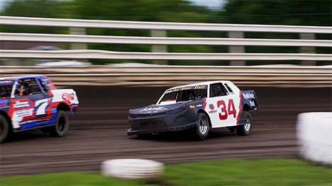 Two race cars zoom around a dirt track. The car on the left (in the lead), is number 7. The car on the right (behind car 7) is number 34.
