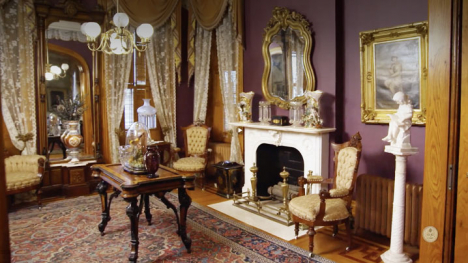 A Victorian-style parlor features ornate chairs, a marble fireplace, a decorative mirror, framed artwork, heavy drapes, and a patterned rug on polished wooden floors.