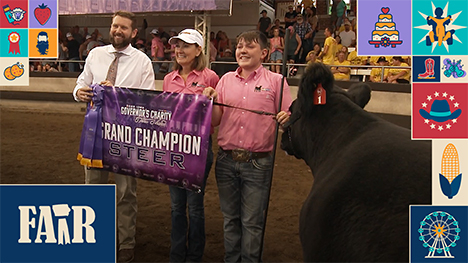 Gov. Kim Reynolds is awarded Grand Champion prize in the Governor's Charity Steer Show. A winning steer and a boy stand to her left and a man stands to her right. They hold purple Grand Champion banner above the dirt floor of the Livestock Pavilion.