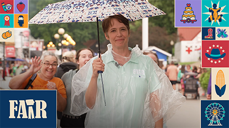 Charity Nebbe holds an umbrella and in the rain at the Iowa State Fair while some women photobomb her in the background with a friendly wave.