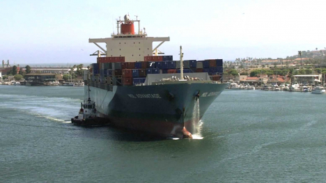 Container ship carrying stacked cargo containers moves through a harbor alongside a tugboat.